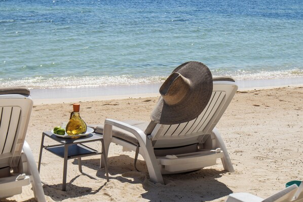 A beach chair with a hat, a bottle, and a plate with food on a table by the sea.