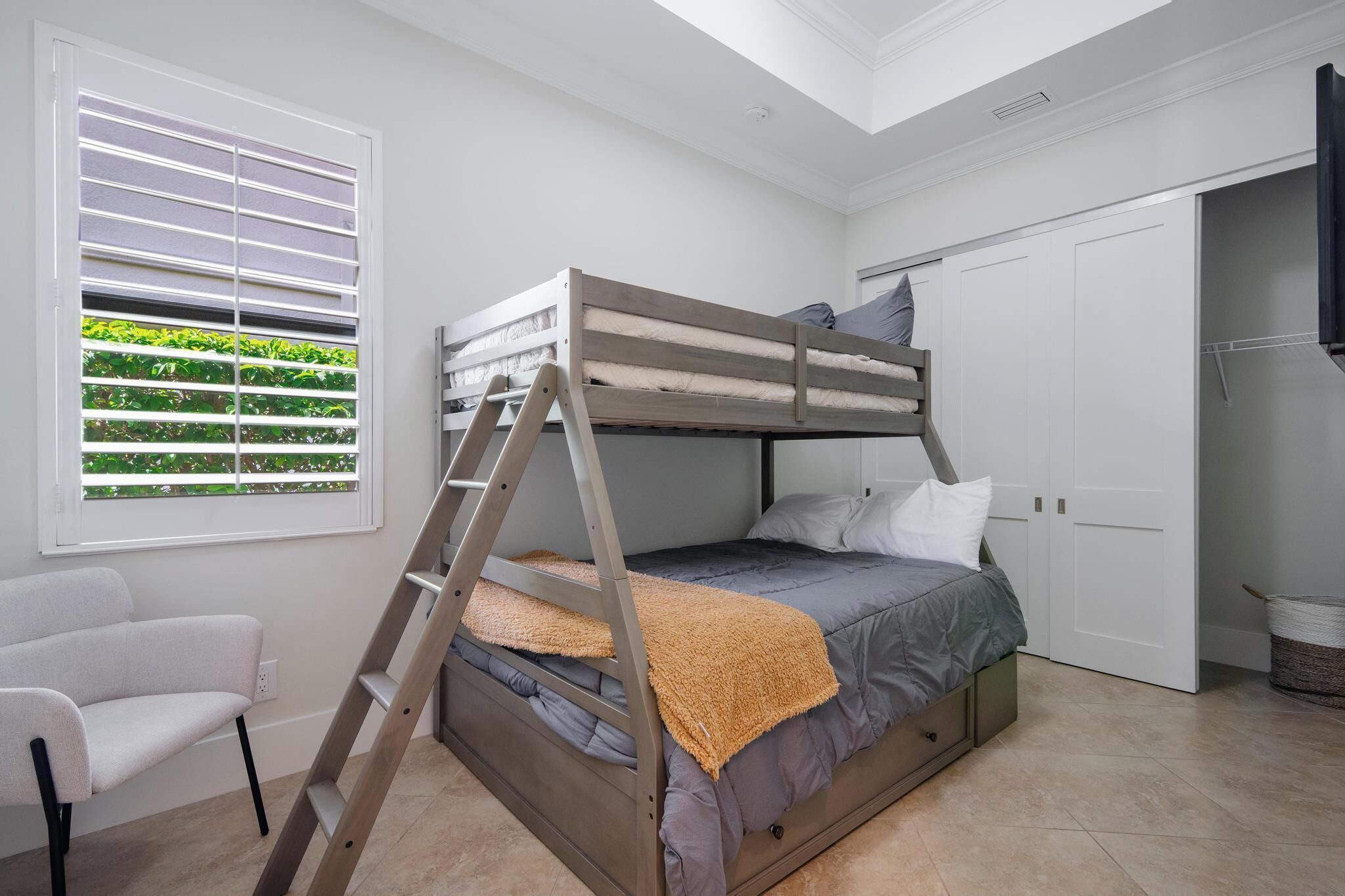 A bunk bed with a ladder, a white chair, a window with blinds, and a door.