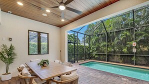 Zona de piscina con mesa y sillas de madera, rodeada por un recinto de cristal con vistas a un paisaje tropical.