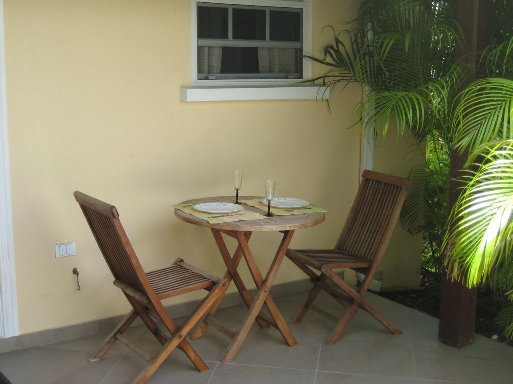 A small patio with a round wooden table and two wooden chairs, a window above, and a potted plant to the right.