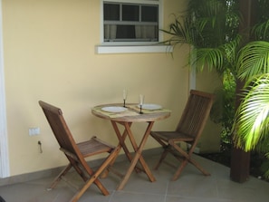 A small patio with a round wooden table and two wooden chairs, a window above, and a potted plant to the right.