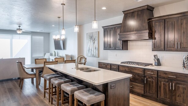 A modern kitchen with dark wood cabinets, a large island with stools, pendant lighting, and a dining area with a view of the outdoors. - Canyon Crest Escape (St. George)