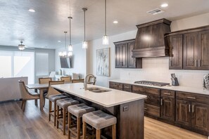 Una cocina moderna con armarios de madera oscura, una isla grande con taburetes, iluminación colgante y un área de comedor con vistas al exterior.
