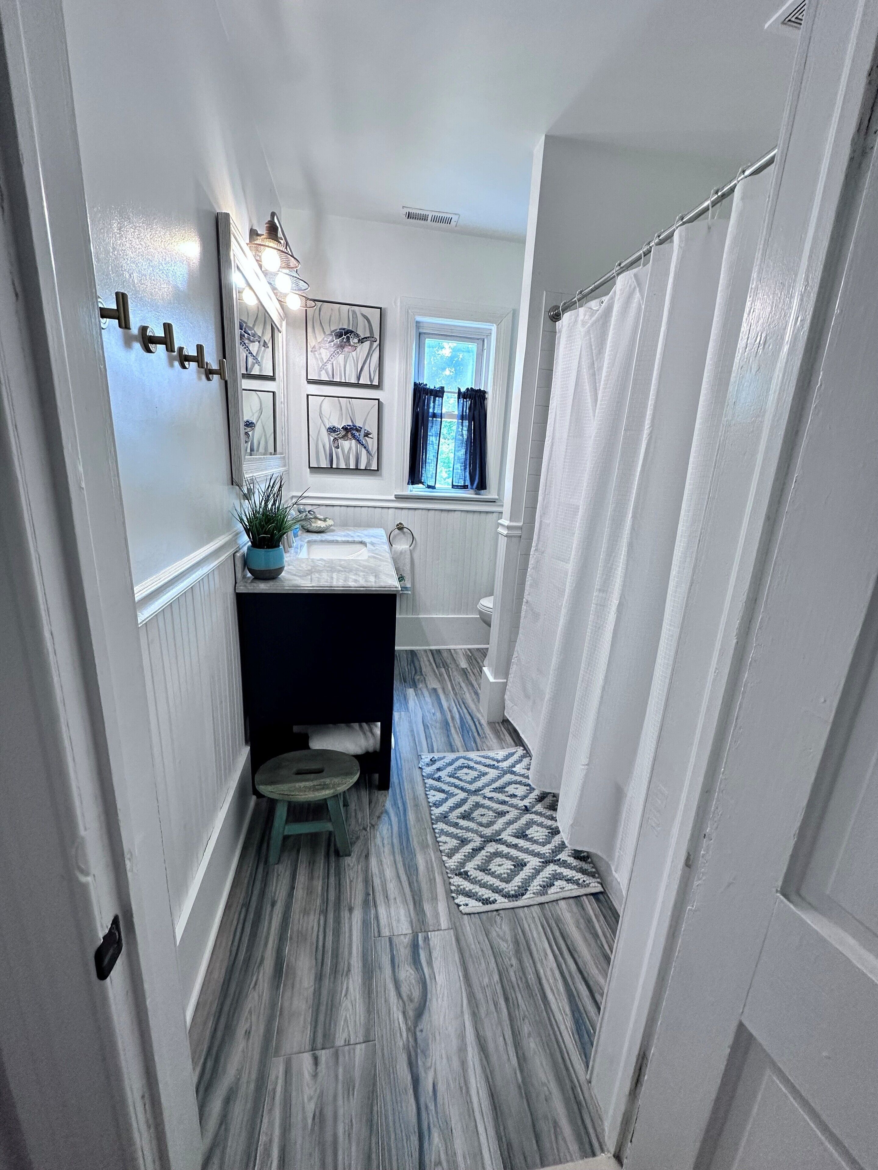 Un couloir étroit donnant sur une salle de bain équipée d’une fenêtre, d’un lavabo et d’un rideau de douche.