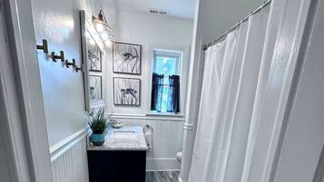 Un couloir étroit donnant sur une salle de bain équipée d’une fenêtre, d’un lavabo et d’un rideau de douche.