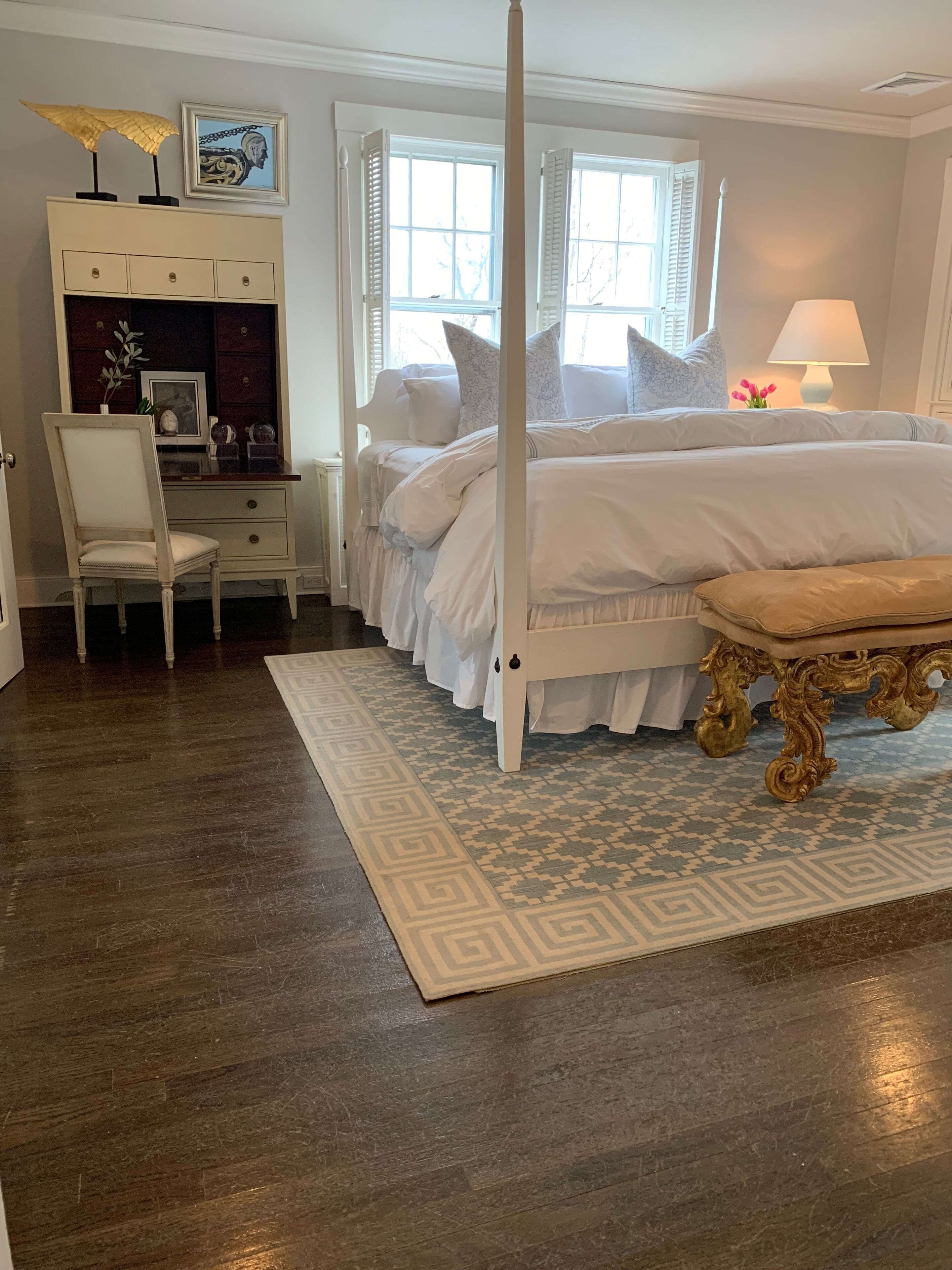 A bedroom with a four-poster bed, a desk with a chair, a vanity with a mirror, and a window with shutters.