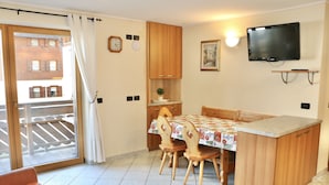 Una cocina pequeña con armarios de madera, una mesa de comedor con sillas, un televisor montado en la pared y un gran ventanal corredizo que da a un espacio exterior.