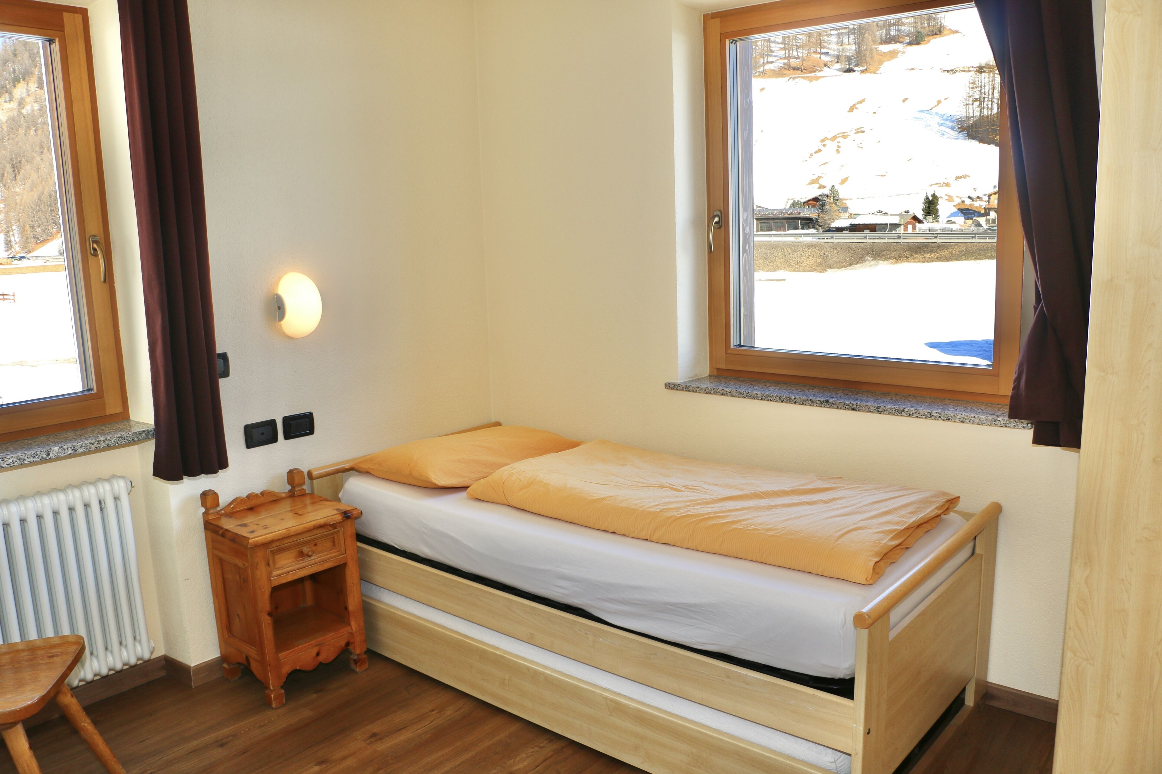 A small, single-bed room with a wooden bed, a nightstand, a wall-mounted light, and a window with a view of a snowy landscape.