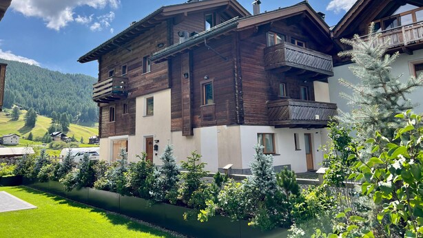 Un edificio de madera de varios pisos con balcones, rodeado por un jardín cuidado y un paisaje montañoso.