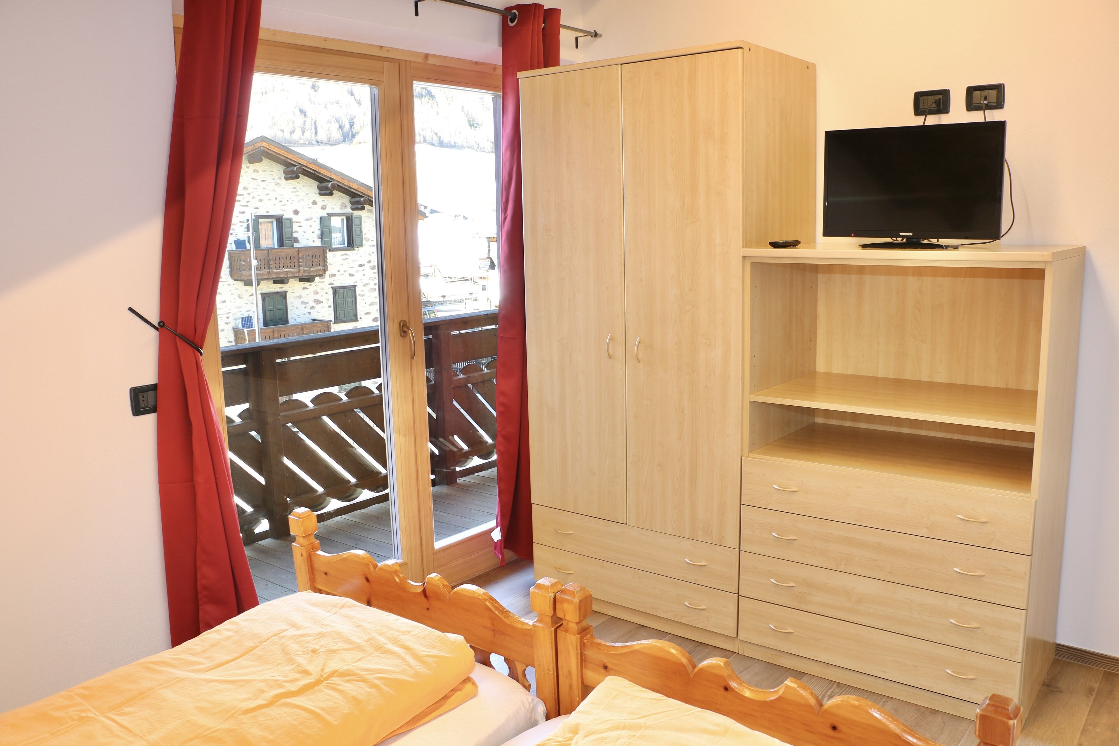 A bedroom with a bed, a wooden wardrobe, a television on a stand, and a balcony with a wooden railing.