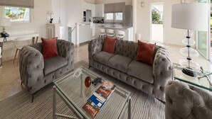 Uma sala de estar espaçosa com um sofá e poltronas cinzas, uma mesa de centro de vidro e uma cozinha moderna ao fundo.