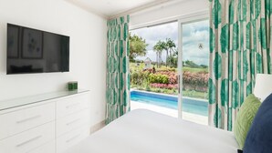 Quarto com uma cama grande, TV montada na parede e vista para uma piscina e jardim.