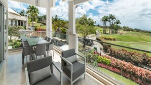 Uma varanda espaçosa com vista para um campo de golfe, área de refeições com mesa e cadeiras, e um design arquitetônico moderno com vigas brancas e guarda-corpos de vidro.