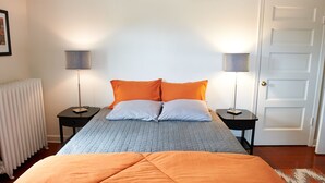 Un dormitorio con cama, dos mesitas de noche, dos lámparas, un radiador y una puerta.
