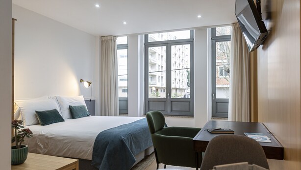 A hotel room with a bed, a desk, a chair, a TV, and large windows with curtains. - Feel Corporate Housing Marquês I (Oporto)