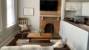 A living room with a brick fireplace, a television mounted above it, a wooden chair, a sofa, and a kitchen area with cabinets and appliances. - Cozy Apartment in the Heart of MainStrasse (Covington)