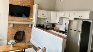 A kitchen with a brick fireplace, white cabinets, a stainless steel refrigerator, and a wooden dining table with a vase of flowers. - Cozy Apartment in the Heart of MainStrasse (Covington)