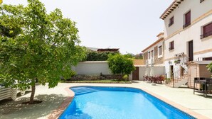 A swimming pool with a curved edge, surrounded by a paved patio. There are outdoor tables and chairs, and a building with a balcony in the background. - Complex of 3 apartments with swimming pool (Melegís)