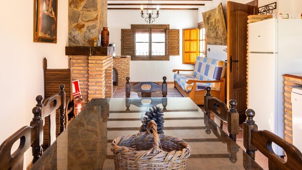 A dining area with a long table, wooden chairs, a stone wall, a wooden ceiling, and a window with shutters. - Complex of 3 apartments with swimming pool (Melegís)