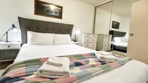 Een net opgemaakt bed met een kleurrijke deken en opgevouwen handdoeken, een bedlamp, een nachtkastje, een dressoir en een grote spiegel.