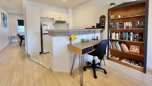 Een moderne keuken met witte kastjes, een ingebouwde oven en een aanrecht. Er is een klein eiland met een spoelbak en een stoel. Een boekenplank vol boeken bevindt zich rechts.