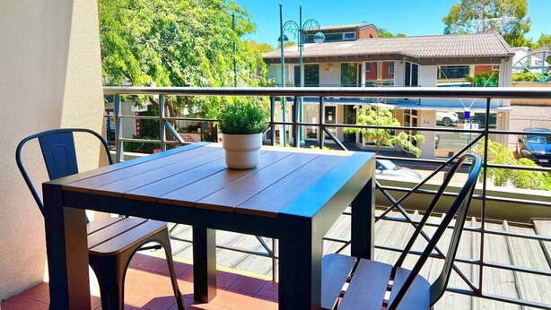 Een balkon met een houten tafel en stoelen, een plant in pot en uitzicht over een woonwijk met huizen en bomen.