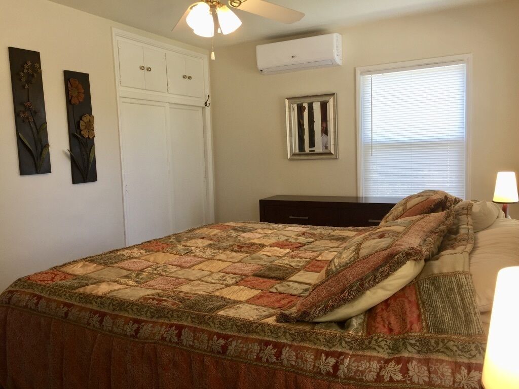 A bedroom with a bed, a wardrobe, a ceiling fan, an air conditioner, a window with blinds, and a nightstand with a lamp.