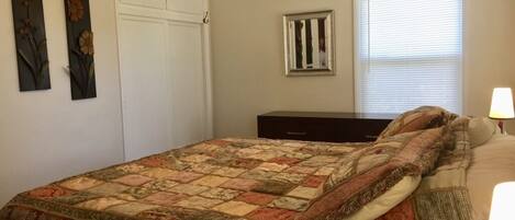 A bedroom with a bed, a wardrobe, a ceiling fan, an air conditioner, a window with blinds, and a nightstand with a lamp.