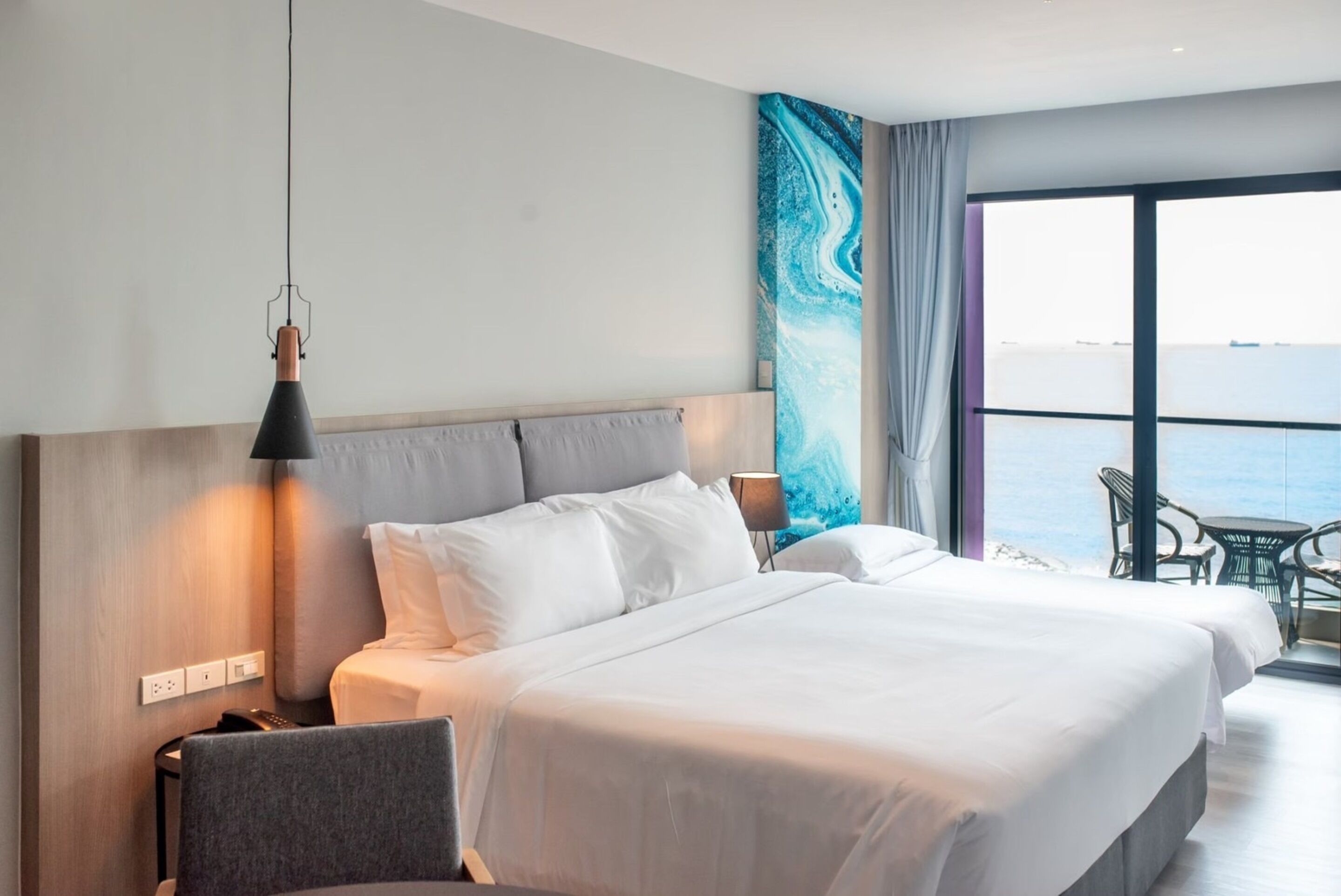 A hotel room with two beds, a large window with a view of the sea, a chair, and a table.