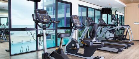 Un gimnasio con varias cintas de correr y bicicletas estáticas, ventanas amplias con vista al mar y suelo de madera.