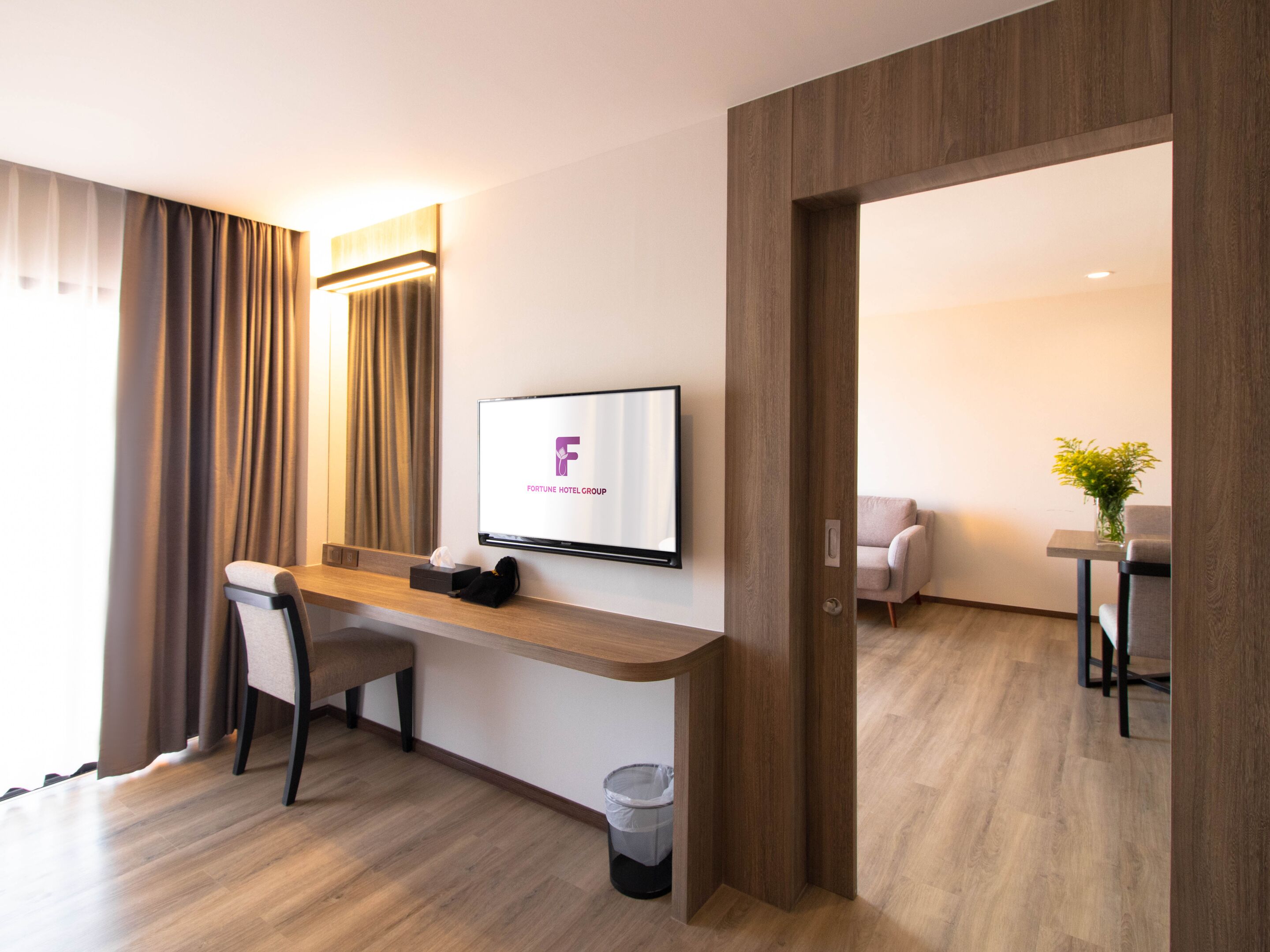 Hotelkamer met een houten bureau, een stoel, een aan de muur bevestigde televisie waarop het logo van Fortune Hotel Group te zien is, een kleine prullenbak en uitzicht op een andere kamer met een bank en een bloemenvaas.