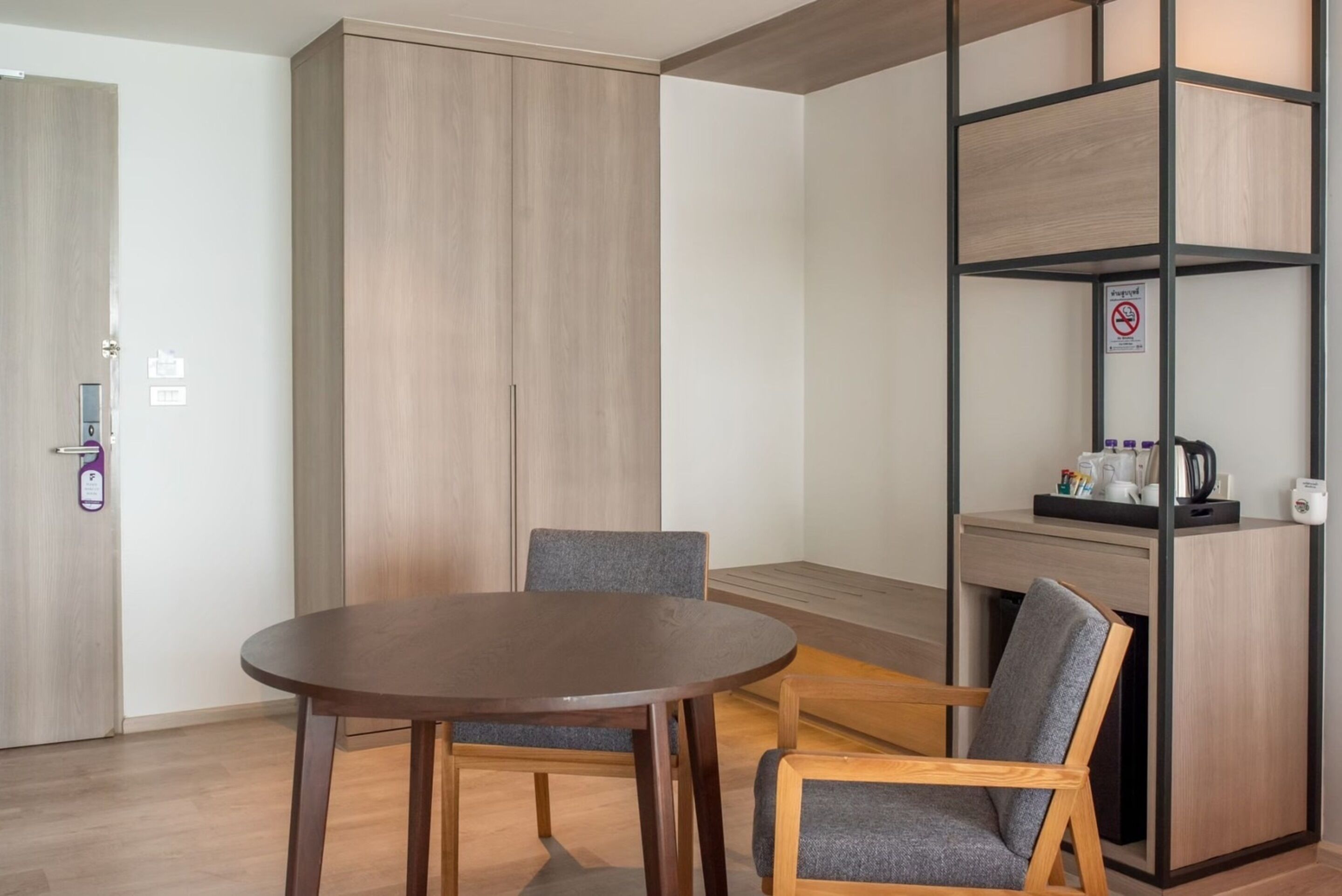 Una habitación moderna con una mesa de madera, dos sillas grises, un armario y un mueble con hervidor y cafetera.