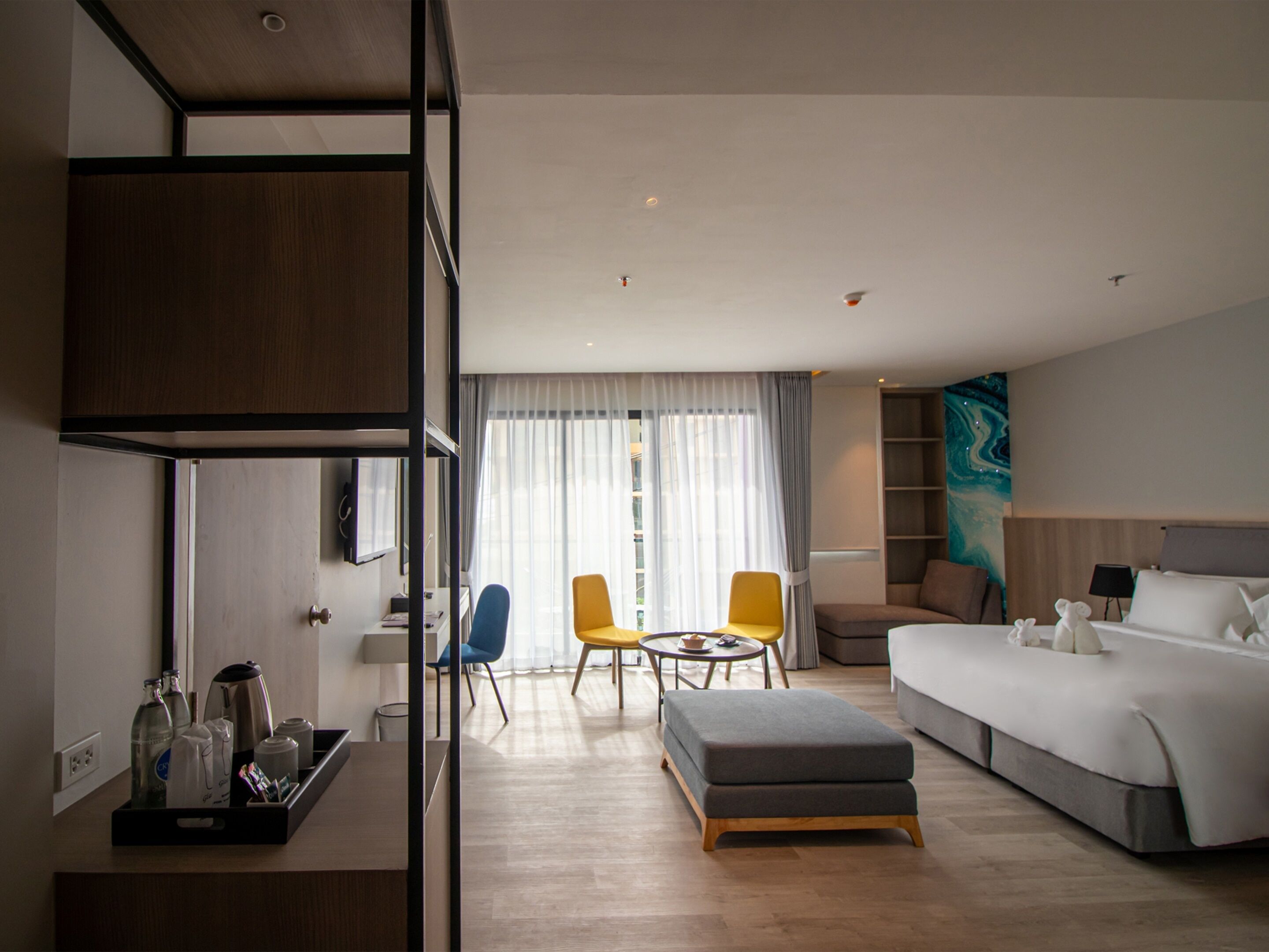 A modern hotel room with a bed, a sofa, a small table, and two chairs. There is a desk with a lamp and a tray with bottles and a kettle.