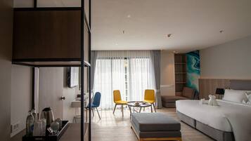 A modern hotel room with a bed, a sofa, a small table, and two chairs. There is a desk with a lamp and a tray with bottles and a kettle.