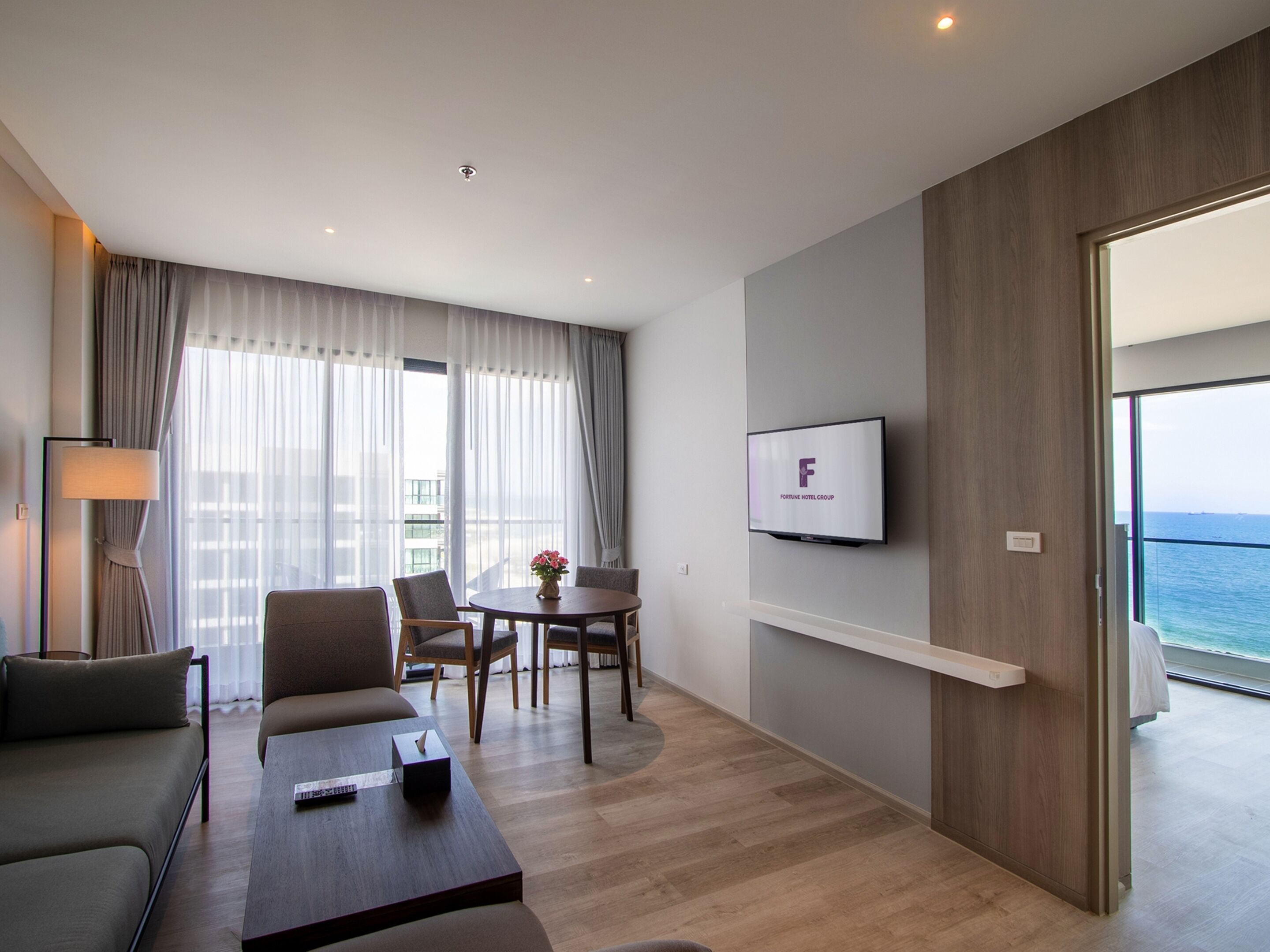 Quarto de hotel moderno com sofá, mesa de centro, televisão na parede, varanda com vista para o mar e área de jantar com cadeiras e mesa.
