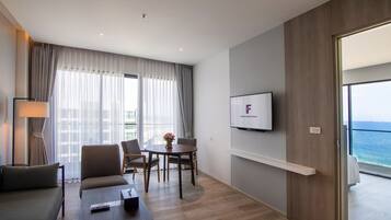 Quarto de hotel moderno com sofá, mesa de centro, televisão na parede, varanda com vista para o mar e área de jantar com cadeiras e mesa.