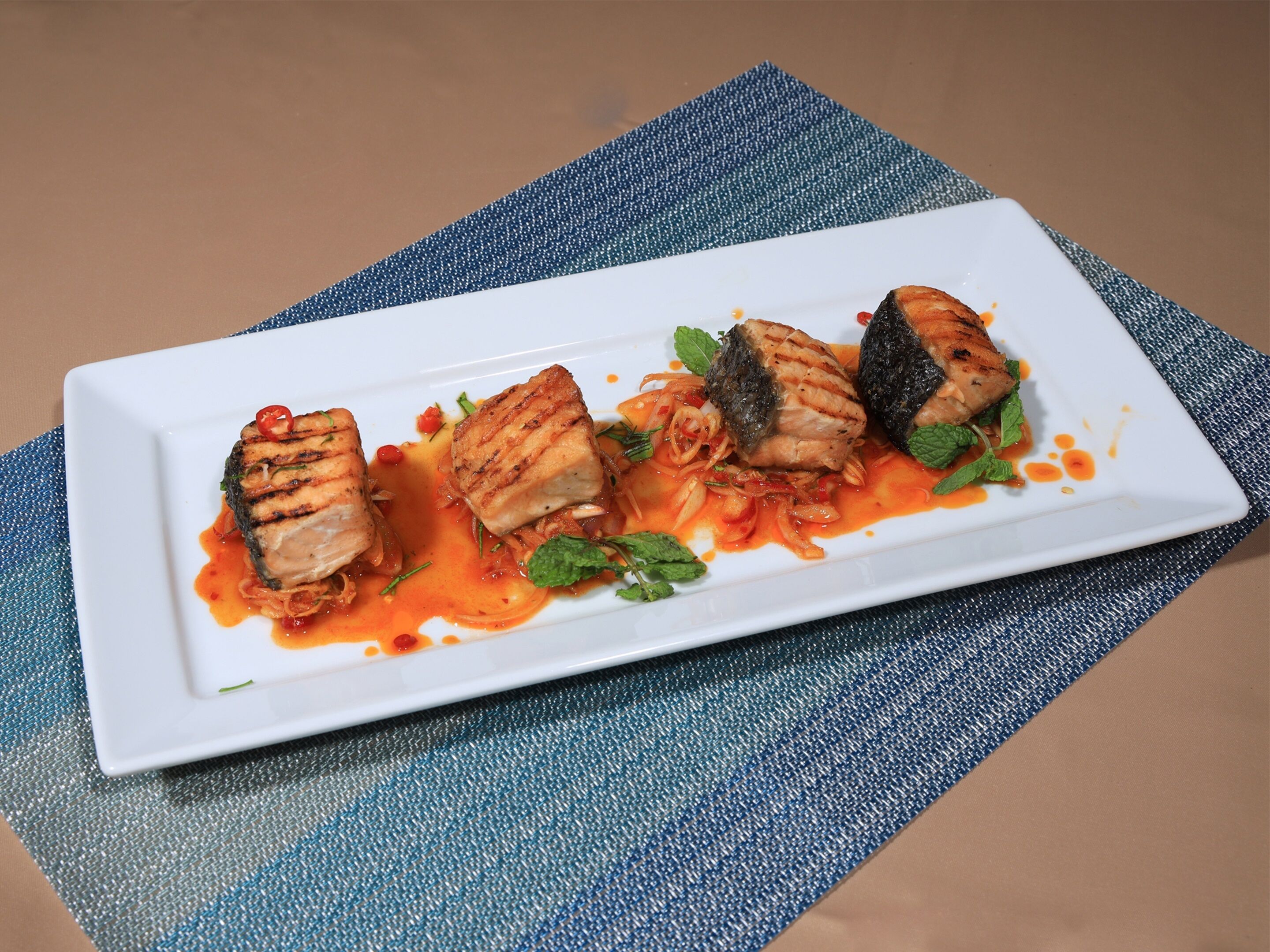 A white rectangular plate with grilled fish and sauce on a textured blue surface.