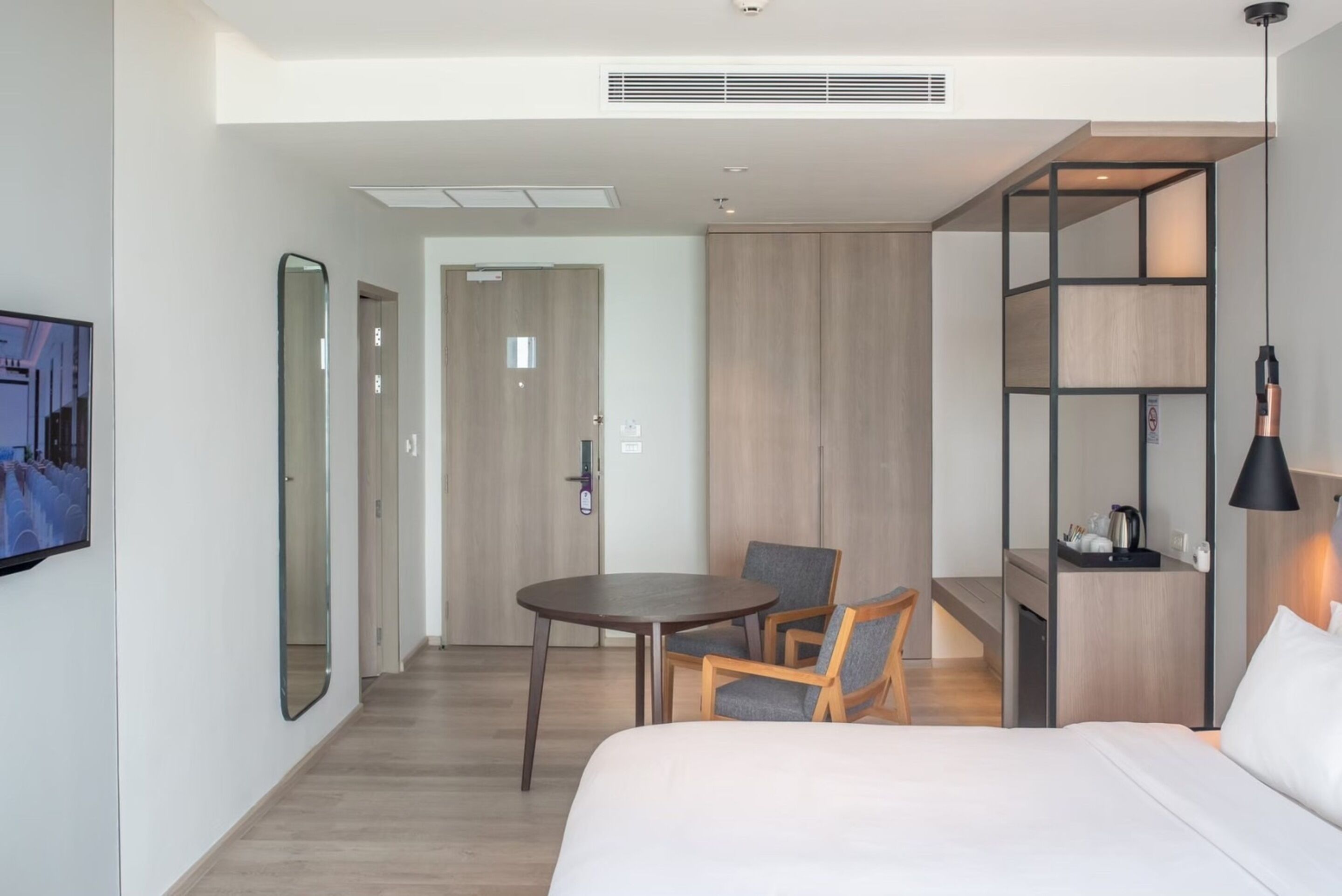 A modern hotel room with a bed, a desk with a chair, a TV, and a large mirror.