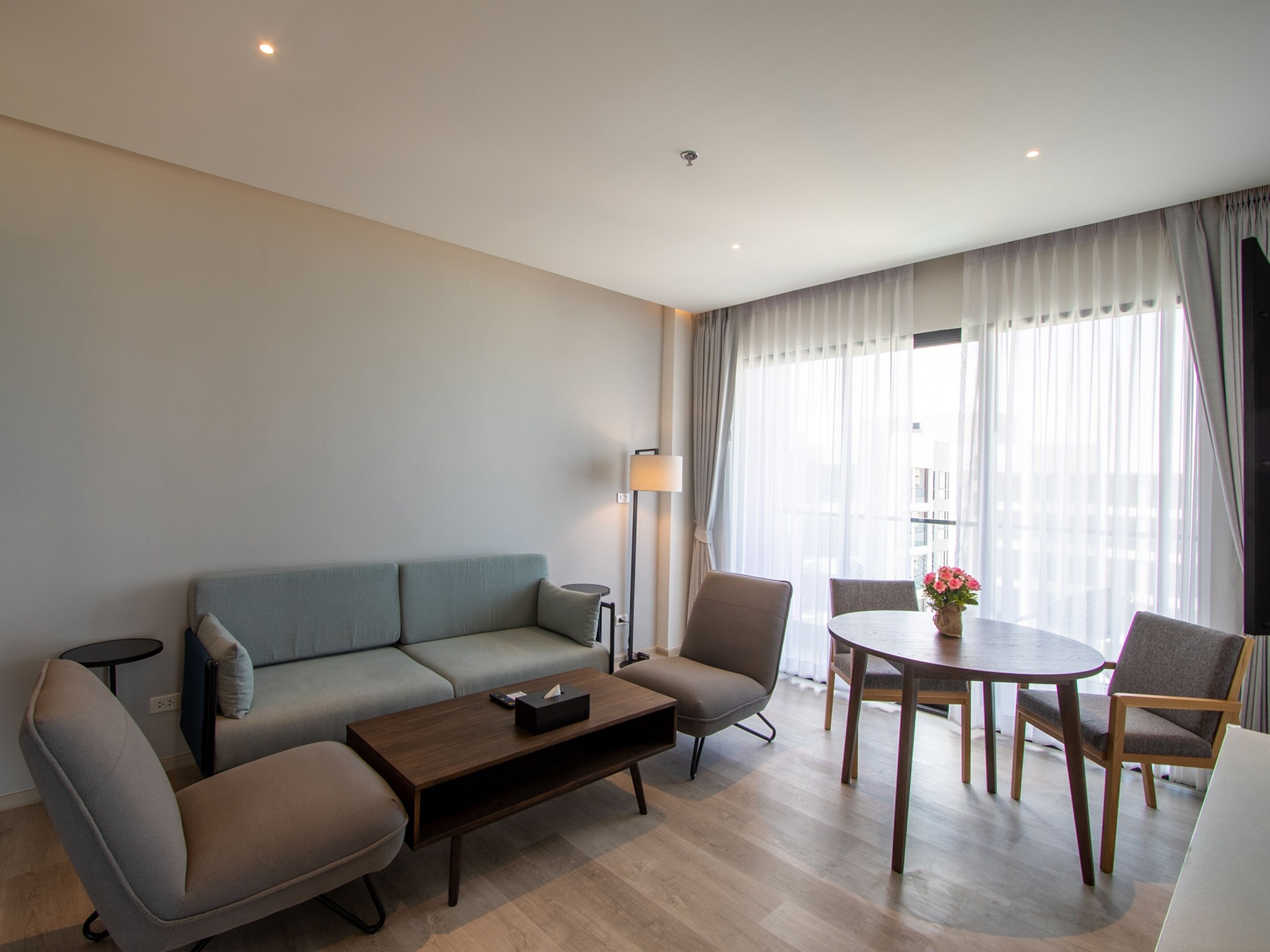 A modern hotel room with a sofa, a coffee table, a dining table with chairs, and a lamp.