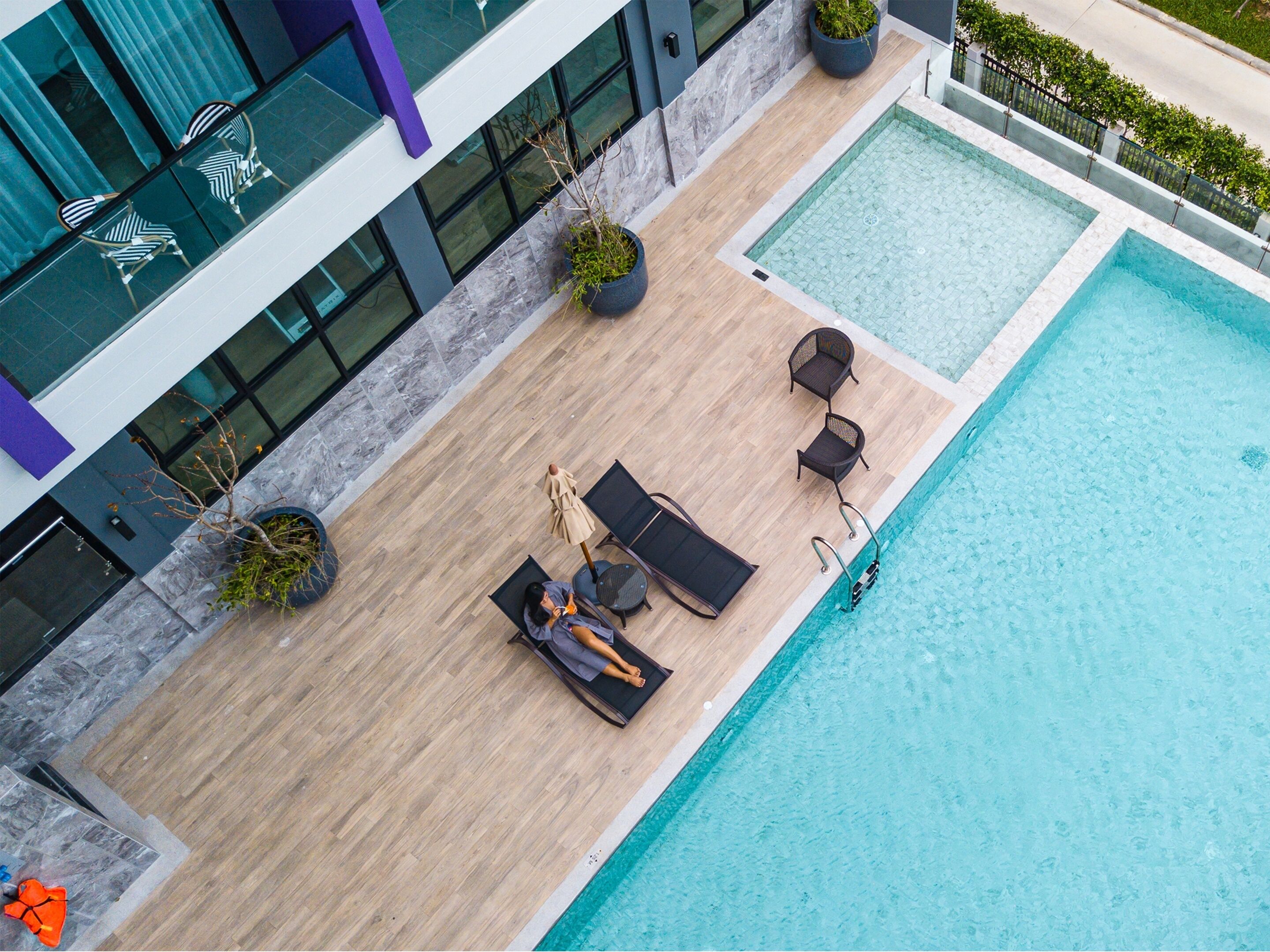 An aerial view of a modern hotel pool area with a wooden deck, lounge chairs, and potted plants.