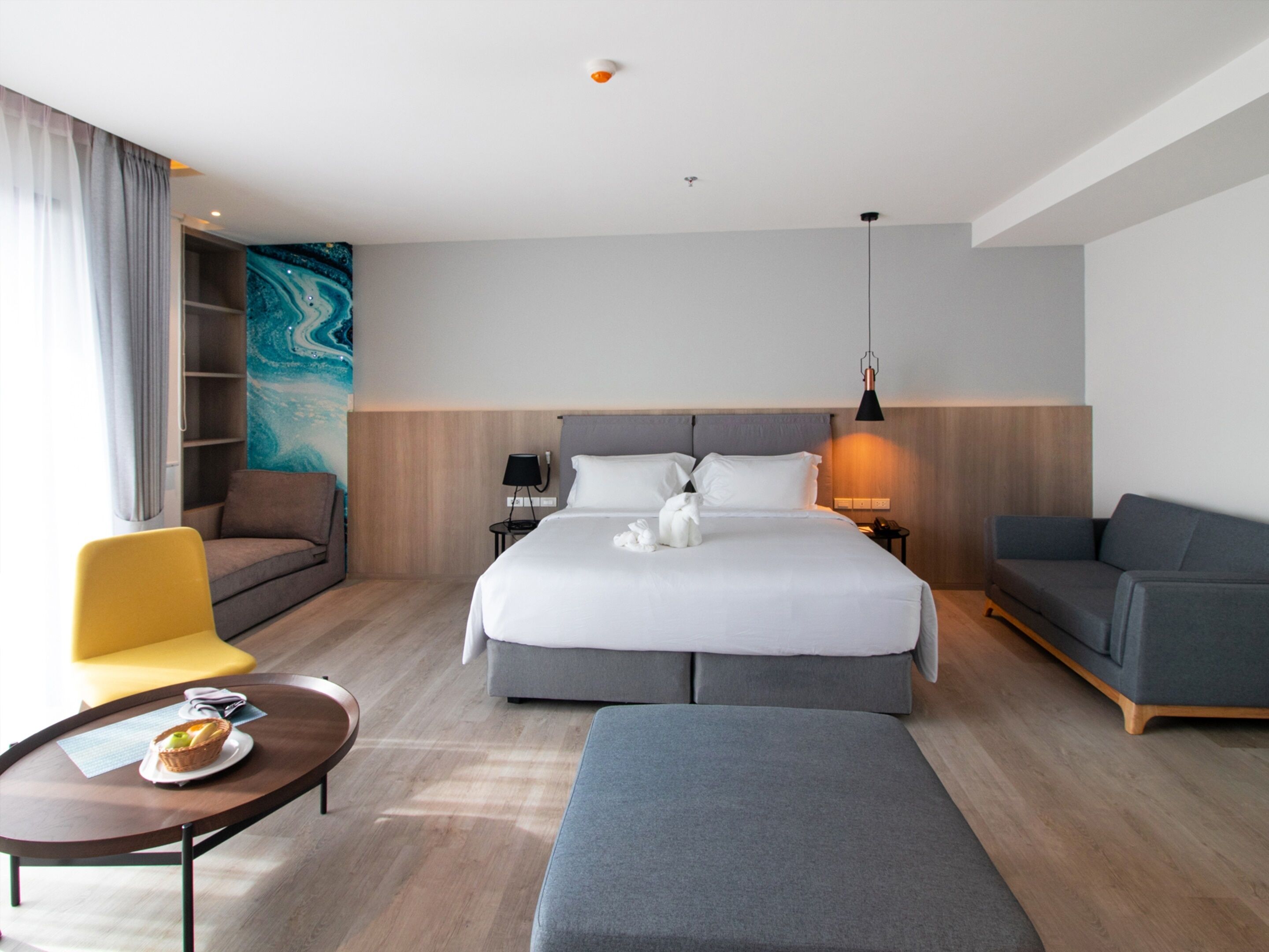 Una habitación de hotel moderna con una cama grande, un sofá, una silla, una mesita con una cesta y una pared con un cuadro abstracto azul.