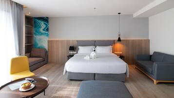 Una habitación de hotel moderna con una cama grande, un sofá, una silla, una mesita con una cesta y una pared con un cuadro abstracto azul.