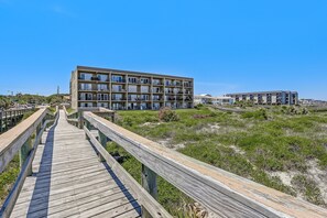 Ein Holzsteg führt zu einem modernen, mehrstöckigen Gebäude mit Balkonen, umgeben von dichter Vegetation.