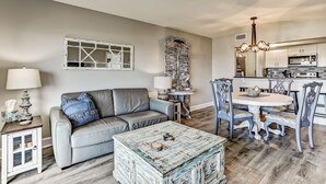 Ein Wohnzimmer mit grauem Sofa, einem Couchtisch und einem Essbereich mit blauen Stühlen und einer Pendelleuchte.