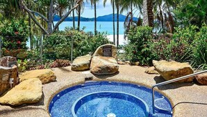 A circular hot tub surrounded by rocks and tropical plants, with a view of the sea and mountains in the background.