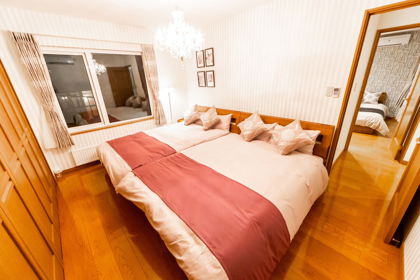 A hotel room with a large bed, wooden flooring, a chandelier, and a window with curtains.
