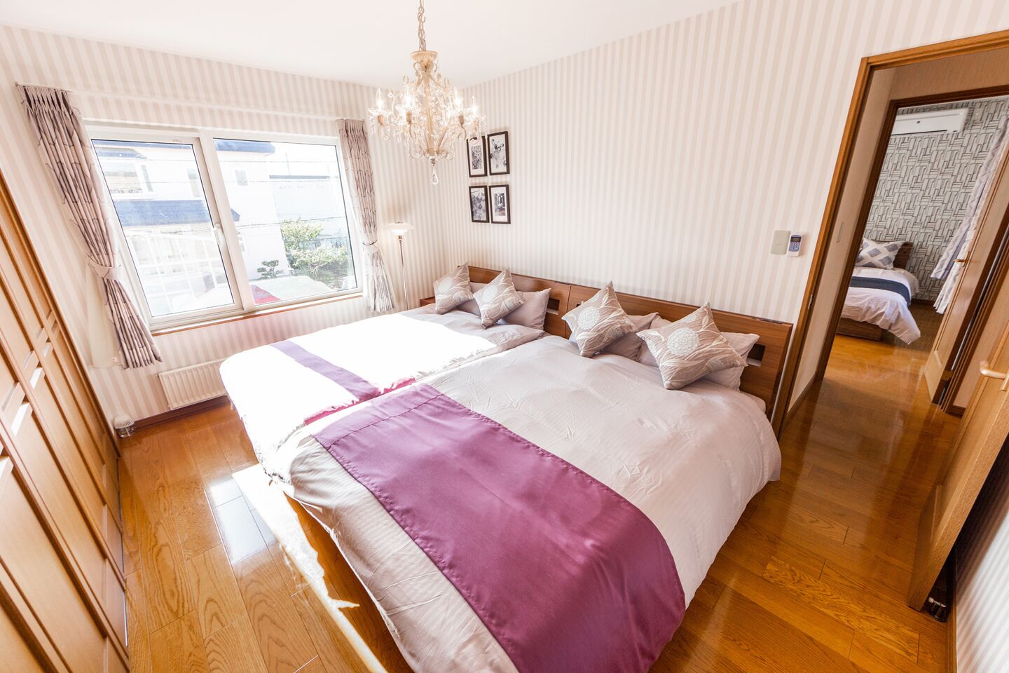 A bedroom with a large bed, wooden flooring, a chandelier, and a window with curtains.