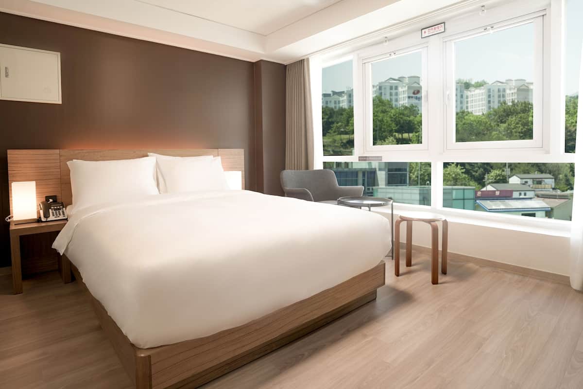 Habitación de hotel moderna con una cama grande, mesita de noche, lámpara y vistas a edificios a través de la ventana.