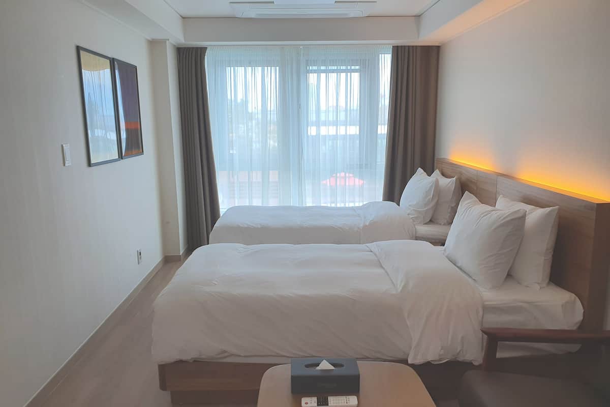 Habitación de hotel con dos camas, una mesita de noche con una caja de pañuelos y vistas a la ciudad a través de amplias ventanas.