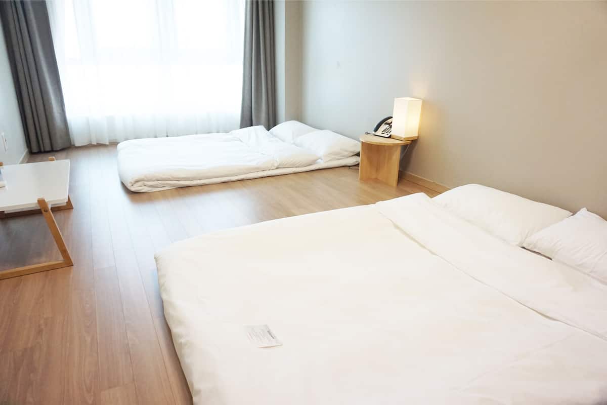 Habitación de hotel con dos camas, una mesita de noche con lámpara, suelo de madera y ventana con cortinas.